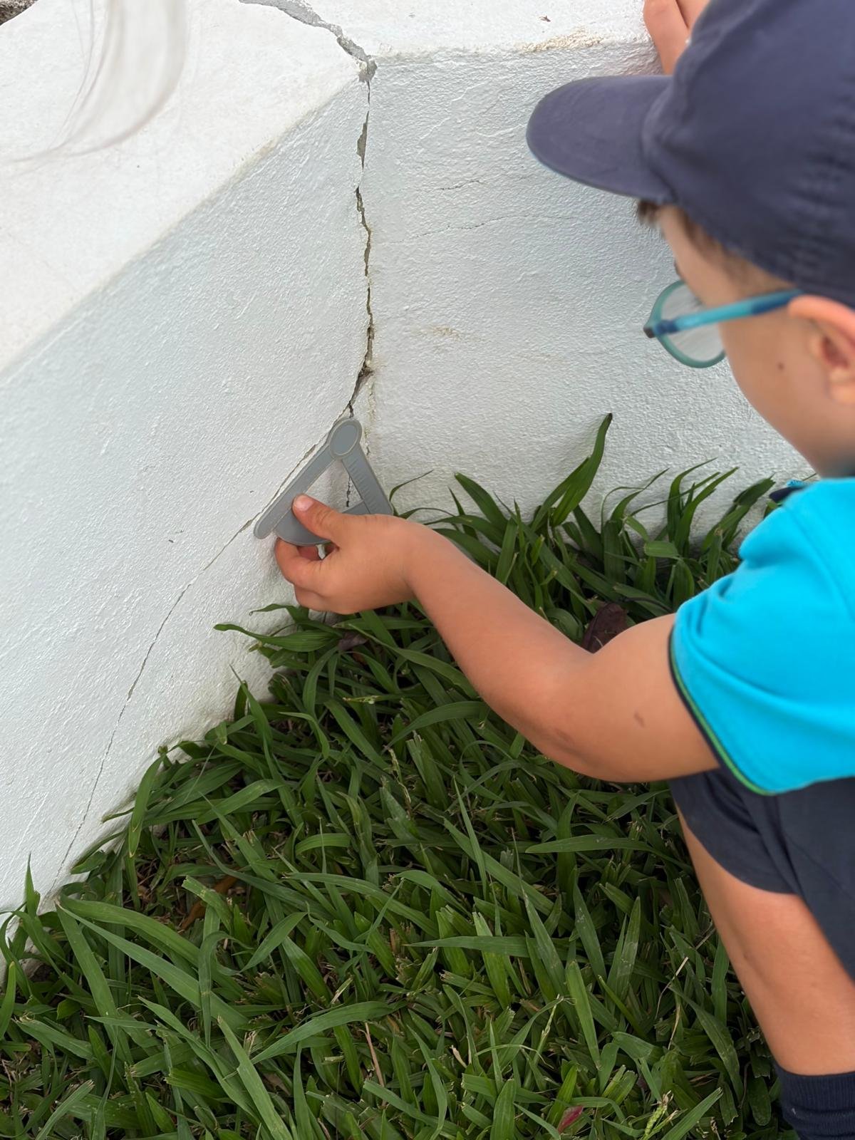 Criança de óculos e boné a encostar o esquadro ao canto de um muro branco, junto à relva